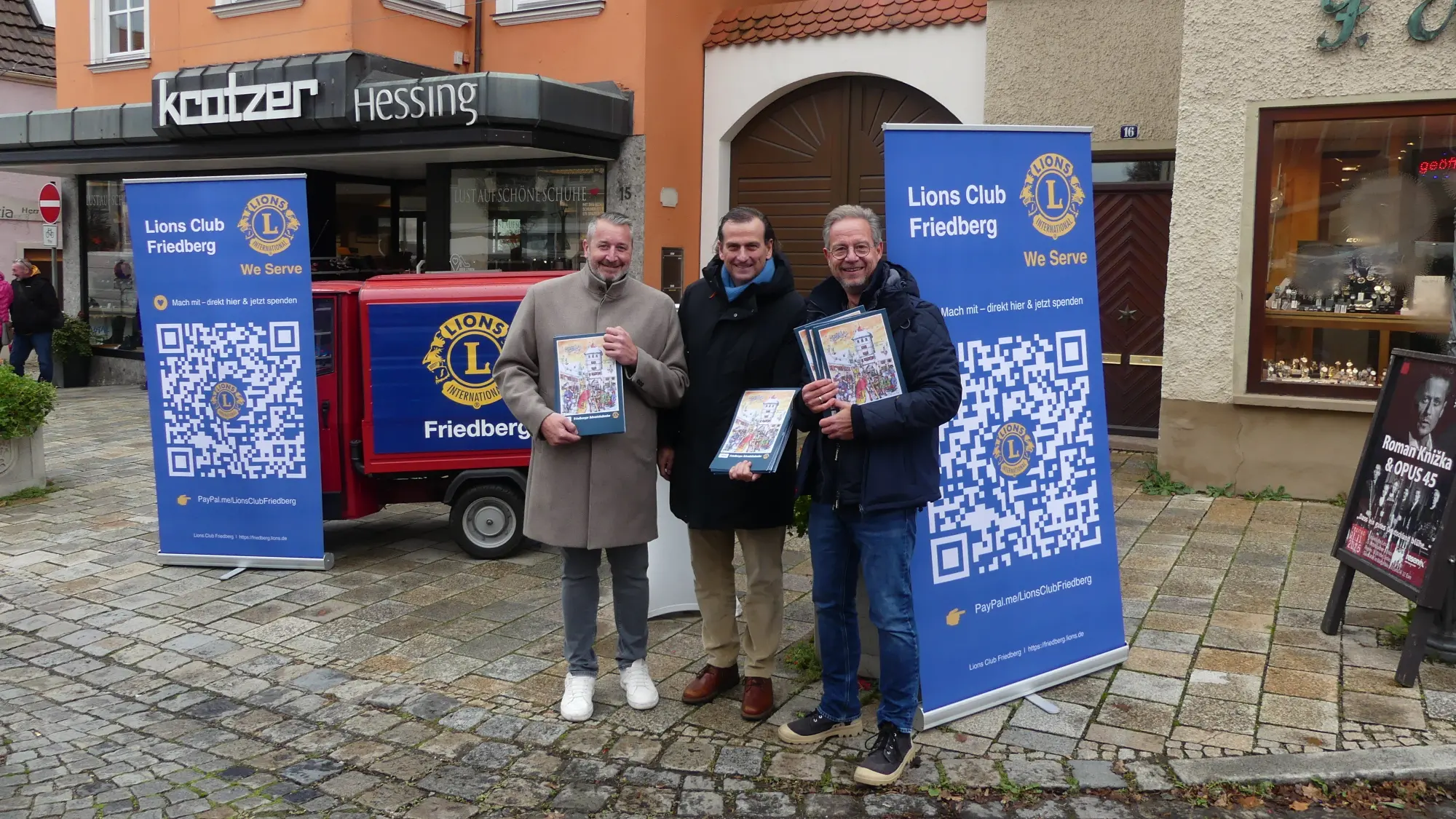 In der Nähe des Rathauses stehen drei Mitglieder des Lions Club Friedbergund deuten auf die baldige Weihnachtszeit hin: Sie verkaufen Adventskalender.